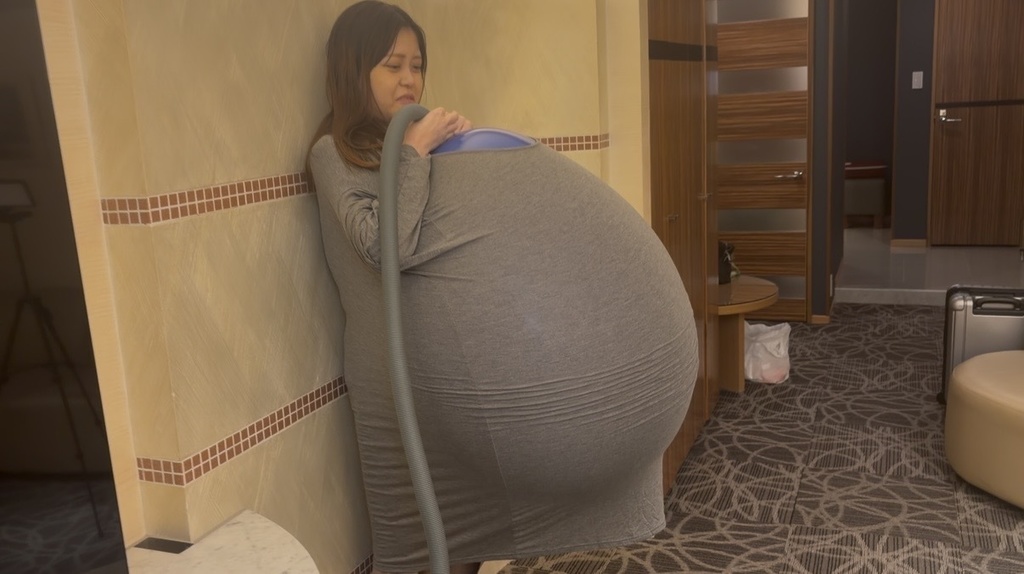 グレーのワンピースの中で風船がふくらみはじめ、壁にもたれるゲストモデルDさん